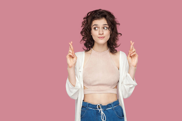 funny hopeful beautiful brunette young woman with curly hairstyle in casual style standing with crossed fingers and looking away on pink background - Zdjęcie, obraz