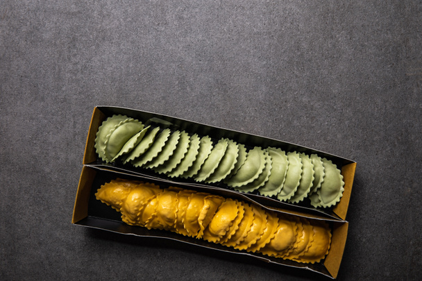 top view of green and yellow raw ravioli in cardboard box on grey textured surface - Фото, зображення