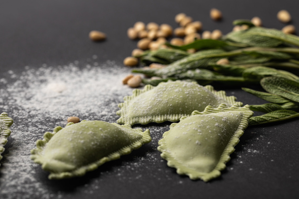 selective focus of green ravioli, sage and scattered flour and pine nuts on black table - Photo, Image