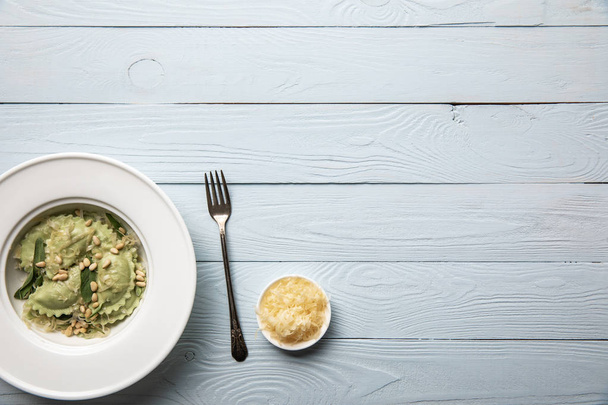 vista superior de ravioles verdes servidos en plato blanco con piñones, salvia y queso rallado sobre mesa de madera blanca
 - Foto, Imagen