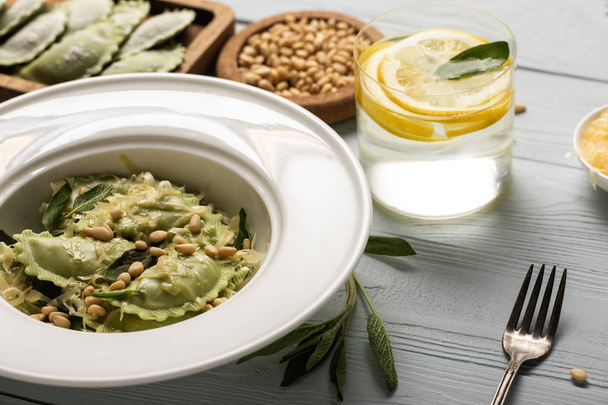 close up view of green ravioli with pine nuts, grated cheese and sage at wooden table near water with lemon - Photo, Image