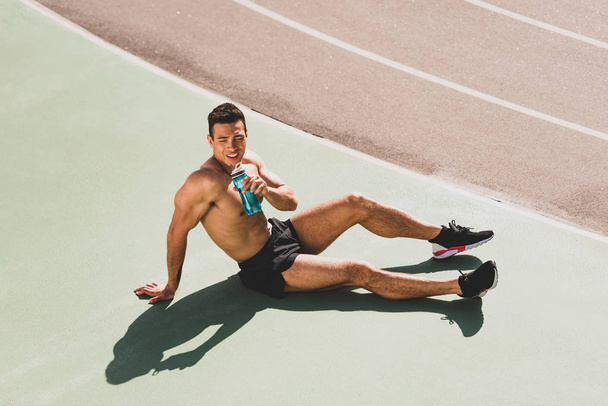 Smějící se smíšený závodní sportovec sedící na stadionu a v lahvi s vodou - Fotografie, Obrázek