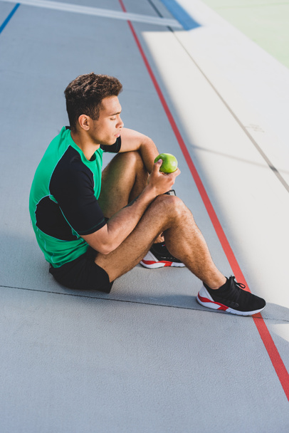 mixed race sportsman sitting on running track and holding green apple - Φωτογραφία, εικόνα