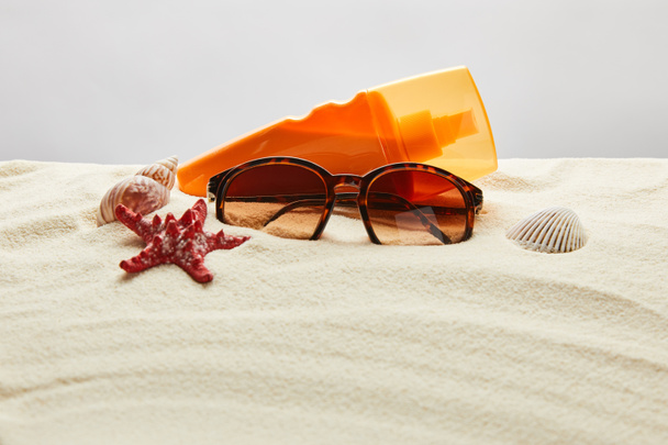 brown stylish sunglasses on sand with red starfish, seashells and sunscreen in orange bottle on grey background - Photo, image