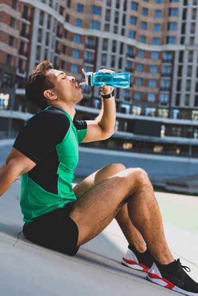 mixed race sportsman sitting on running track at stadium and drinking water from blue bottle  - Foto, Bild