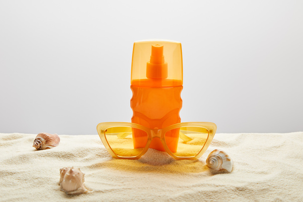 κίτρινα γυαλιά ηλίου και αντηλιακό σε πορτοκαλί μπουκάλι στην άμμο με κοχύλια σε γκρι φόντο - Φωτογραφία, εικόνα
