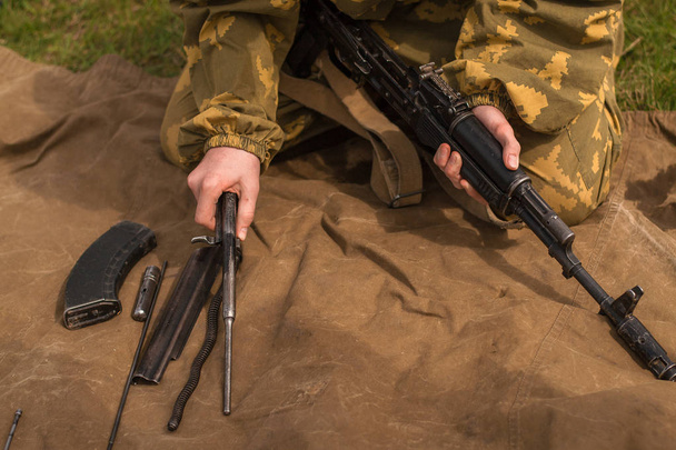 Kamuflaj üniformalı genç bir asker bir silahı söküyor. Bir adam askeri tatbikatlarda çimenlere kalaşnikof saldırı tüfeği topluyor.. - Fotoğraf, Görsel