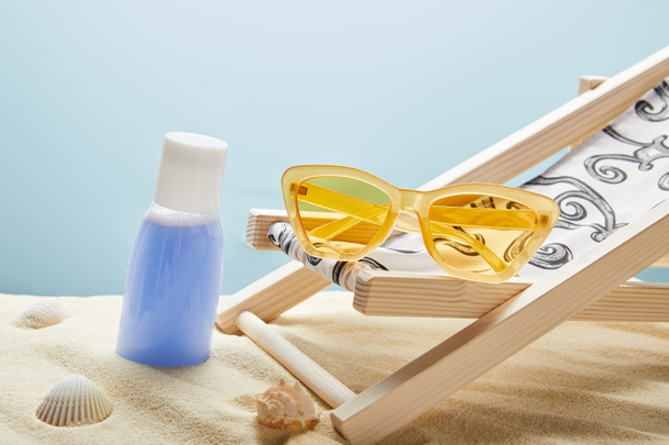 blaue Lotion im Sand in der Nähe von Muscheln, gelbe Sonnenbrille und Liegestuhl auf blauem Hintergrund - Foto, Bild