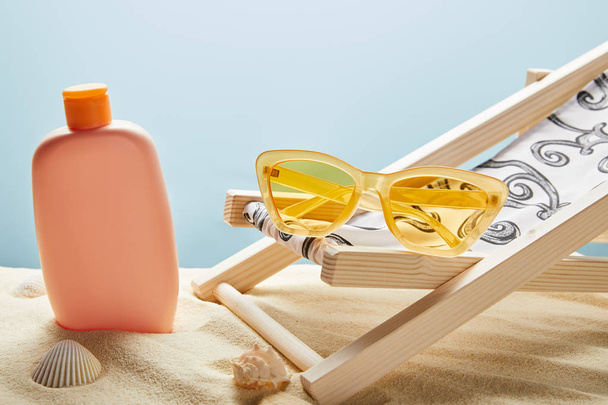 sunscreen lotion in sand near seashells, yellow sunglasses and deck chair on blue background - Photo, Image
