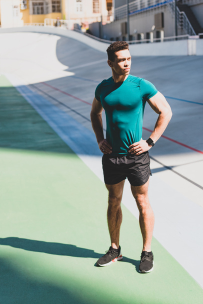 Мускулистый спортсмен смешанной расы, стоящий с руками на бедрах на стадионе при солнечном свете
  - Фото, изображение