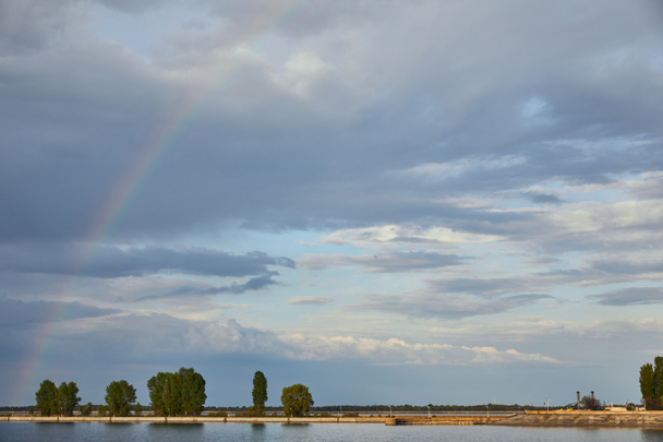 пейзаж голубого солнечного неба, радуга, река и побережье с деревьями
 - Фото, изображение