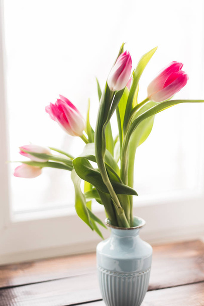 pink tulips in a vase by the bright window on a dark wooden windowsill. spring time - Фото, изображение