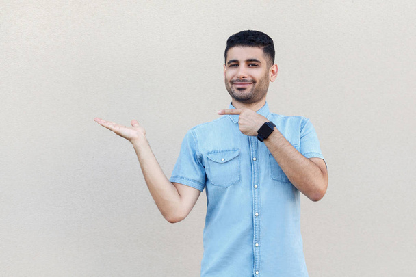happy satisfied handsome young bearded man in blue shirt pointing at empty hand and looking at camera with smile on light beige wall background - Φωτογραφία, εικόνα