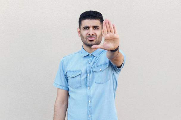 angry serious handsome young bearded man in blue shirt standing with stop hand sign gesture and looking at camera on light beige wall background - 写真・画像