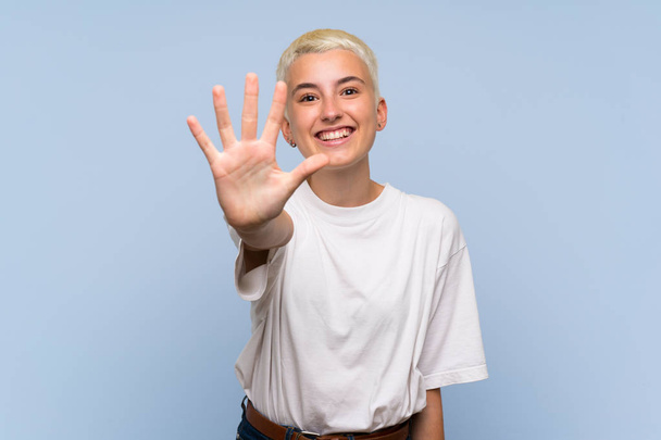 Teenager-Mädchen mit weißen kurzen Haaren über blauer Wand, die mit den Fingern fünf zählt - Foto, Bild