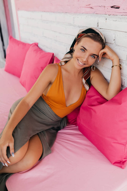 Retrato de chica caucásica riendo en traje de baño naranja se sienta relajadamente con café sobre almohadas rosadas en la terraza de verano
 - Foto, Imagen