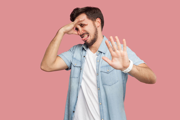 confused handsome bearded young man in blue casual style shirt pinching his nose and showing stop gesture hand sign on pink background - Фото, изображение