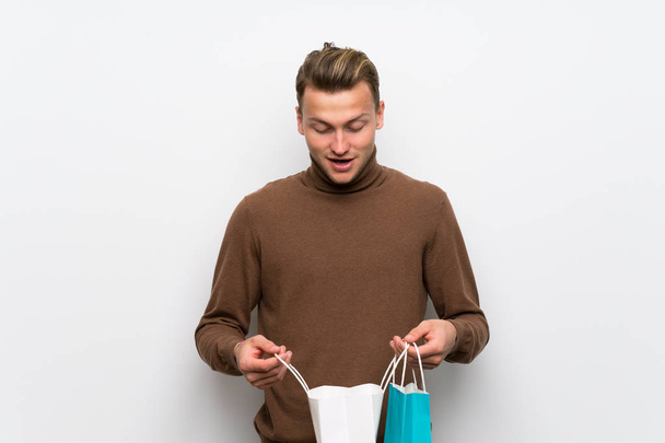 Blonder Mann über isolierte weiße Wand überrascht, während er viele Einkaufstüten in der Hand hält - Foto, Bild