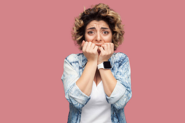 nervous young woman with curly hairstyle in casual blue shirt biting her nails and looking at camera with panic face expression on pink background - Фото, изображение