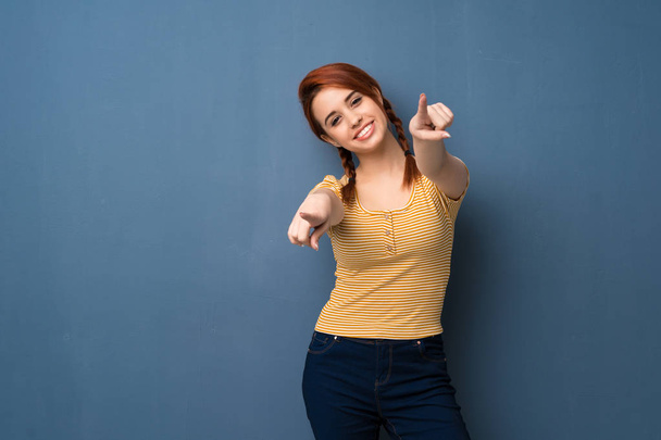 Nuori punapää nainen sinisellä taustalla osoittaa sormella sinua hymyillen
 - Valokuva, kuva