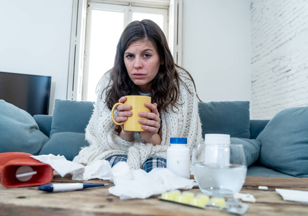 Kranke junge Frau, die auf dem Sofa sitzt und Heißgetränk trinkt, kalte Grippe Medizin, die sich unwohl fühlt, mit Nasenschmerzen und hohen Temperaturen, die nicht zur Arbeit gehen können. Gesundheitsversorgung und medizinisches Konzept. - Foto, Bild