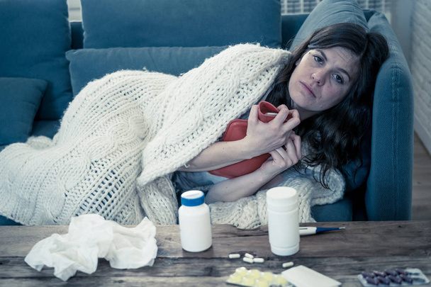 Sick attractive woman with hot water bottle lying on sofa at home feeling unwell with headache sore nose high temperature not being able to go to work. Col flu and Illness in Health Care concept. - 写真・画像