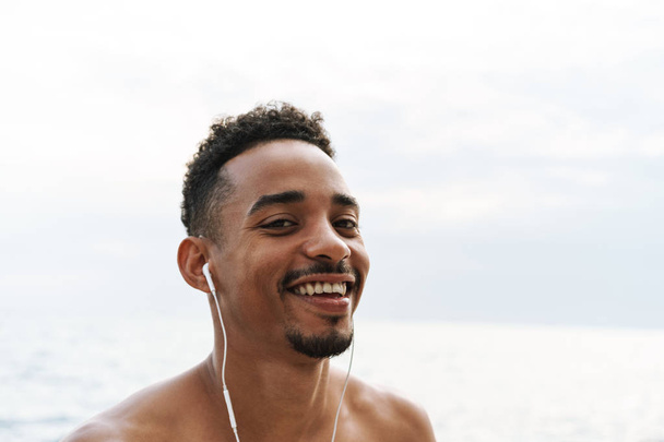 Happy handsome young african sports man outdoors at the beach sea walking listening music with earphones. - Fotó, kép