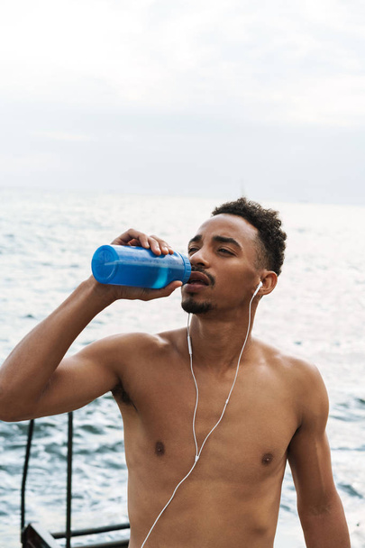 Strong handsome young african sports man outdoors at the beach sea drinking water listening music with earphones. - Fotoğraf, Görsel