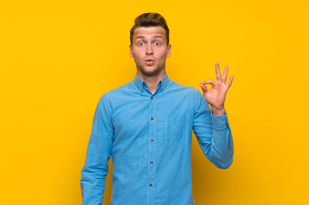 Blonde man over isolated yellow wall surprised and showing ok sign - Photo, Image