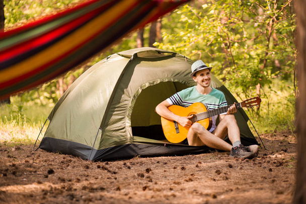 Ευτυχισμένος νεαρός άνδρας κάμπινγκ και strum κιθάρα ορχηστρική μουσική για να χαλαρώσετε με φόντο το ηλιοβασίλεμα του δάσους. Wilderness Ταξιδιωτικός τρόπος ζωής. - Φωτογραφία, εικόνα