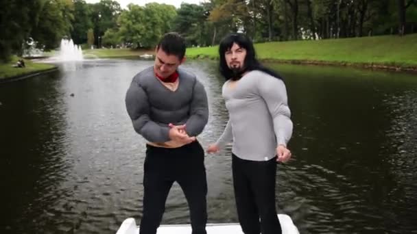 Zwei attraktive Männer in falscher Muskelbrust und armgepolsterten Kostümen stehen im Boot - Filmmaterial, Video