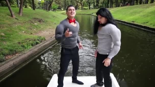 Dois homens engraçados atraentes vestidos com fantasias musculares falsas cantam e dançam em barco
 - Filmagem, Vídeo