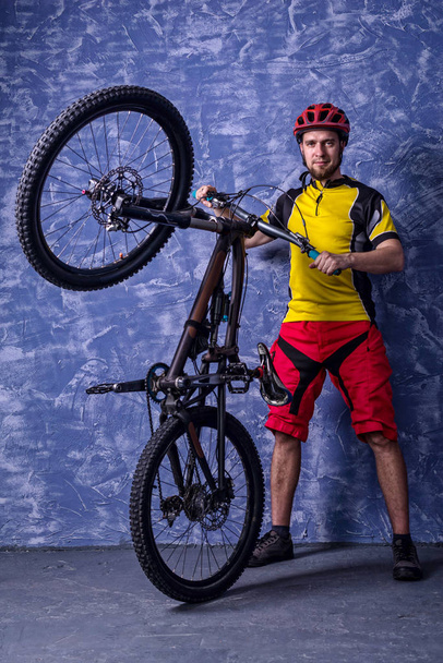 Велосипедист у жовтій футболці та червоних шортах тримає гірський велосипед на тлі синьої стіни
. - Фото, зображення