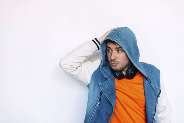 young handsome man wearing jeans jacket with hood on and headphones on his neck standing against white wall while posing on camera - Foto, immagini