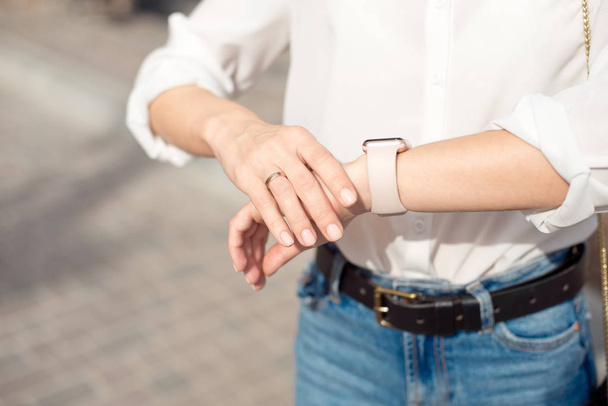 Woman wearing white shirt wearing blue jeans and a gentle nude manicure looks at the watch close-up. - Fotó, kép