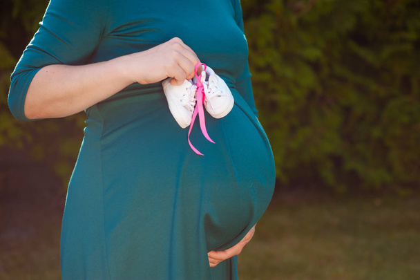 Kis cipő a meg nem született babának a terhes nő hasában. Terhes nő kezében kis baba cipő pihentető otthon a hálószobában. Kis cipő a meg nem született babának a terhes nő hasában - Fotó, kép