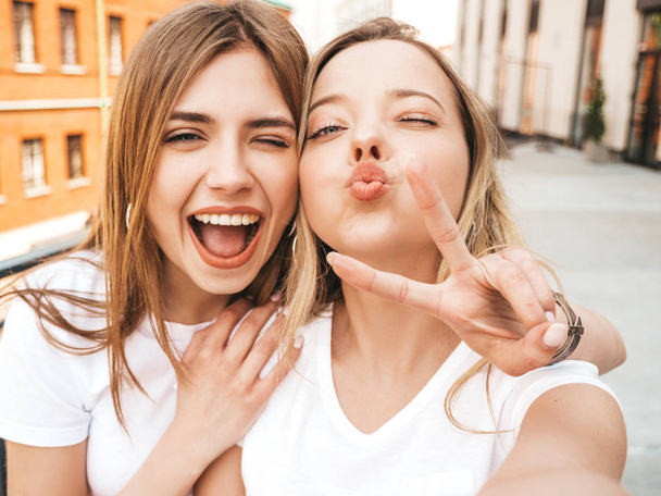 Two young smiling hipster blond women in summer clothes. Girls taking selfie self portrait photos on smartphone.Models posing on street.Female making duck face and shows peace sign - Foto, Imagen