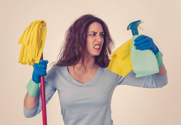 Beautiful angry and upset woman holding mop and cleaning spray feeling frustrated and furious in domestic duties concept and gender stereotypes. Studio portrait isolated on blue background. - Foto, immagini