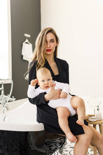 young beautiful blonde girl in stylish classic clothes of black color sits in a bathroom with a baby in a white apartment - Фото, изображение