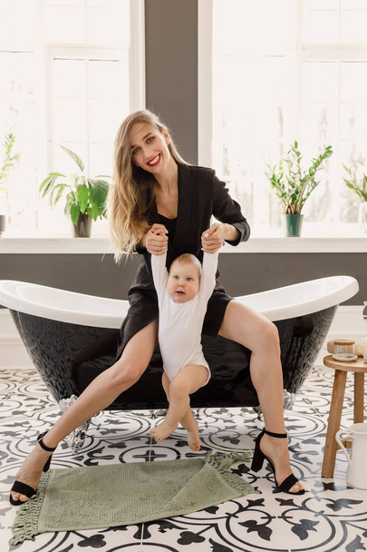 νέα όμορφη ξανθιά κορίτσι στο κομψό κλασικό ρούχα του μαύρου χρώματος κάθεται σε ένα μπάνιο με ένα μωρό σε ένα λευκό διαμέρισμα - Φωτογραφία, εικόνα