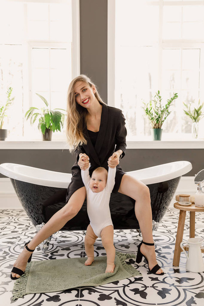 nuori kaunis blondi tyttö tyylikäs klassinen vaatteita musta väri istuu kylpyhuoneessa vauvan kanssa valkoisessa asunnossa
 - Valokuva, kuva