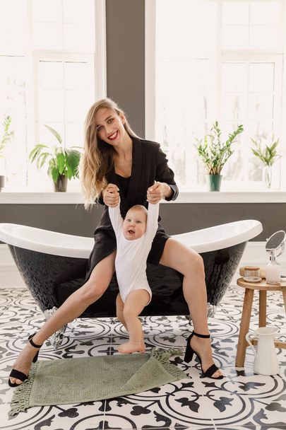 joven hermosa chica rubia en ropa clásica con estilo de color negro se sienta en un baño con un bebé en un apartamento blanco
 - Foto, imagen