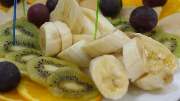 Banai, sinaasappelen, druiven, kiwi gesneden, close-up. Verse fruitmand op een feestelijke eettafel. Geassorteerde gesneden Fruitspiesjes op een plaat. - Video
