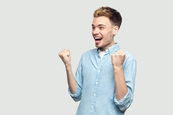 happy surprised handsome young man in light blue shirt standing and celebrating his victory while screaming on grey background - Foto, Imagen