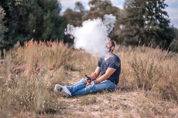 Fumatore barbuto soffia un sacco di fumo utilizzando vaporizzatore sigaretta elettronica in natura. Sigaretta elettronica
. - Foto, immagini