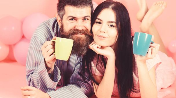 Man and woman on smiling faces lay, pink background. Couple relax in morning with coffee. Couple in love drink coffee in bed. Family tradition concept. Man and woman in domestic clothes, pajamas - Photo, image