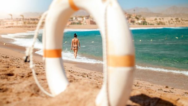 Image de regarder à travers l'anneau de sauvetage en plastique blanc sur jeune homme sexy en petit bikini noir marchant sur la plage
. - Photo, image