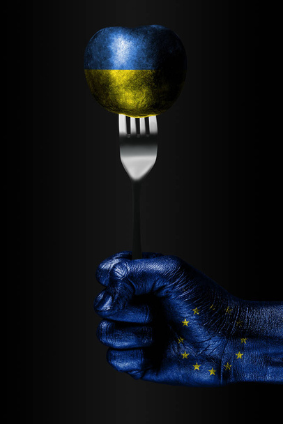 Une main avec un drapeau de l'UE dessiné tient une fourchette, sur laquelle se trouve une balle avec un drapeau de l'Ukraine dessiné, un signe d'influence, de pression, d'adhérence et d'anecxie
. - Photo, image