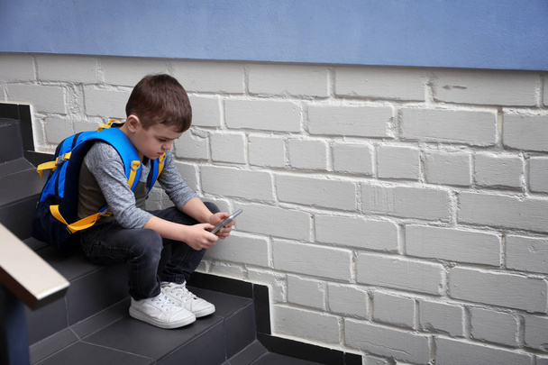 Sad little boy with mobile phone sitting on stairs indoors - Фото, изображение