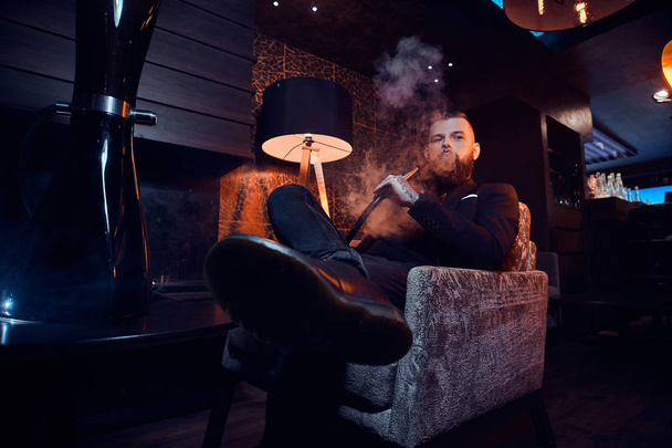 Pensive tattooed man is relaxing on the armchair and is smoking hookah, making hazy vapour - 写真・画像